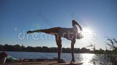 站在瑜伽馆前的年轻人的剪影，摆在湖边的木制码头上。 在大自然训练运动的家伙。 运动员在做